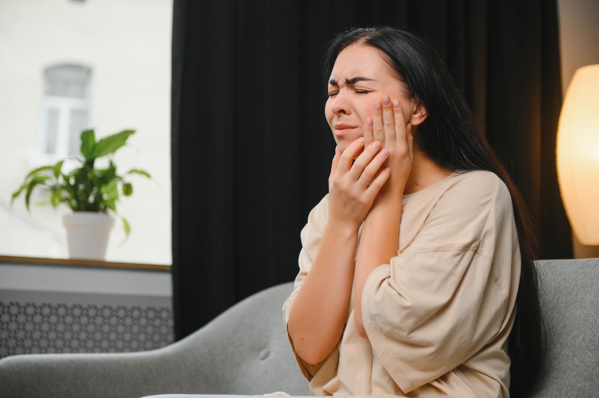 Woman with severe toothache touching her swollen cheek. Young girl feeling discomfort after tooth extraction. Upset lady with inflamed tooth nerve or terrible dental pain sitting on sofa at home