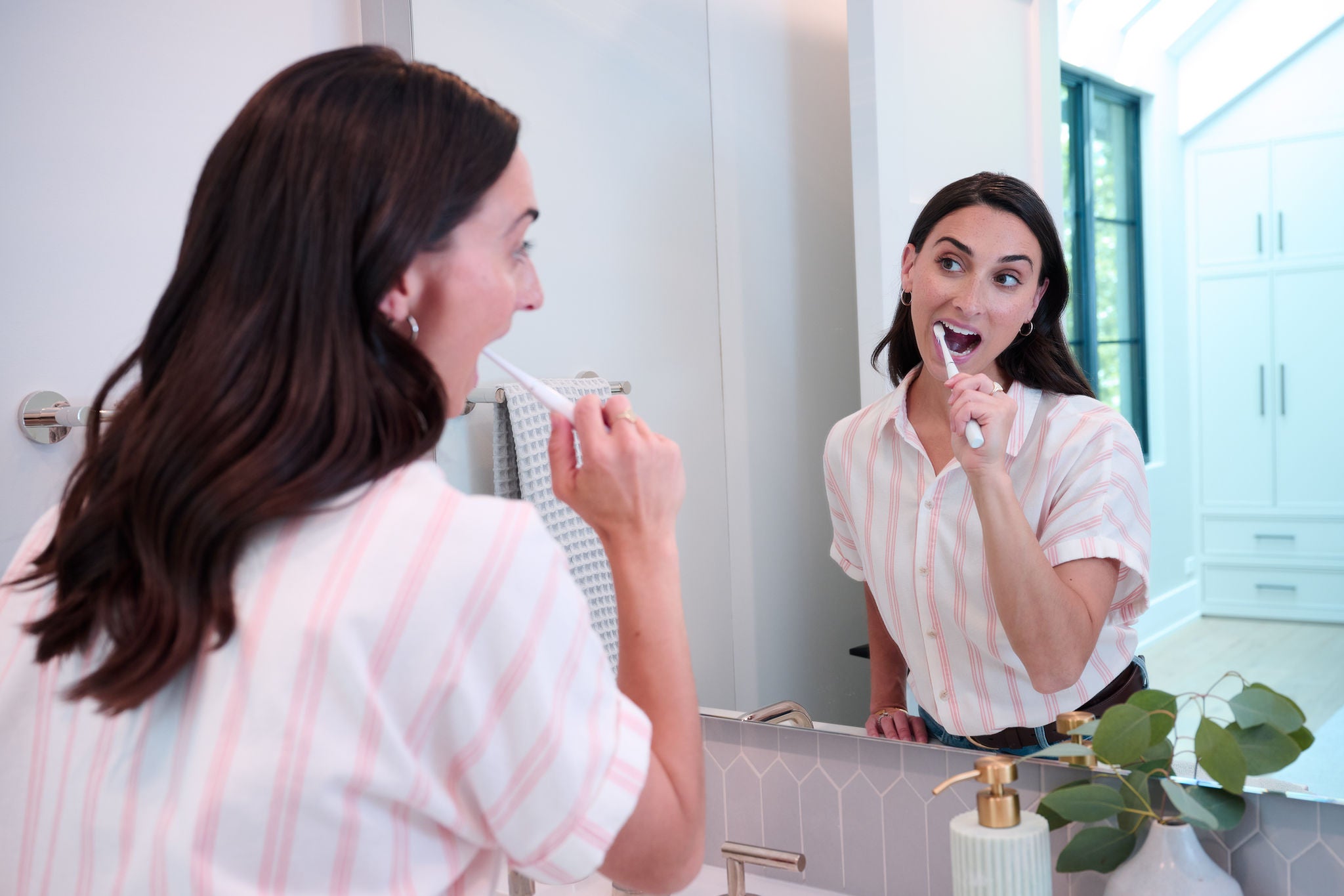 A woman brushes her teeth with a white sonic toothbrush in a modern bathroom. Image resolution: 2048x1365.