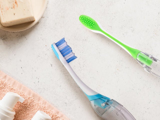 Two GUM TARVEL toothbrushes in blue and green colours shown from the side and back, placed in a natural environment