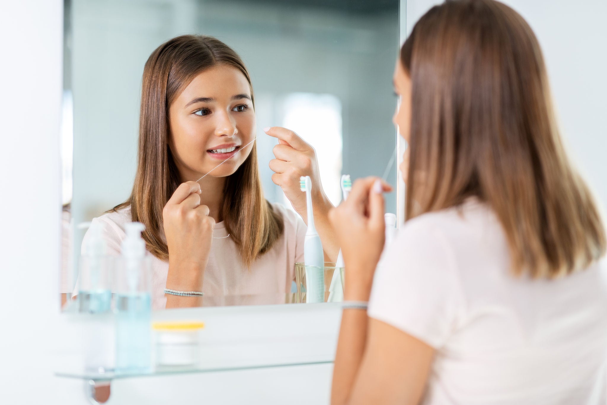 dental care, hygiene and people concept - happy smiling teenage girl with floss cleaning teeth and looking in mirror at bathroom
