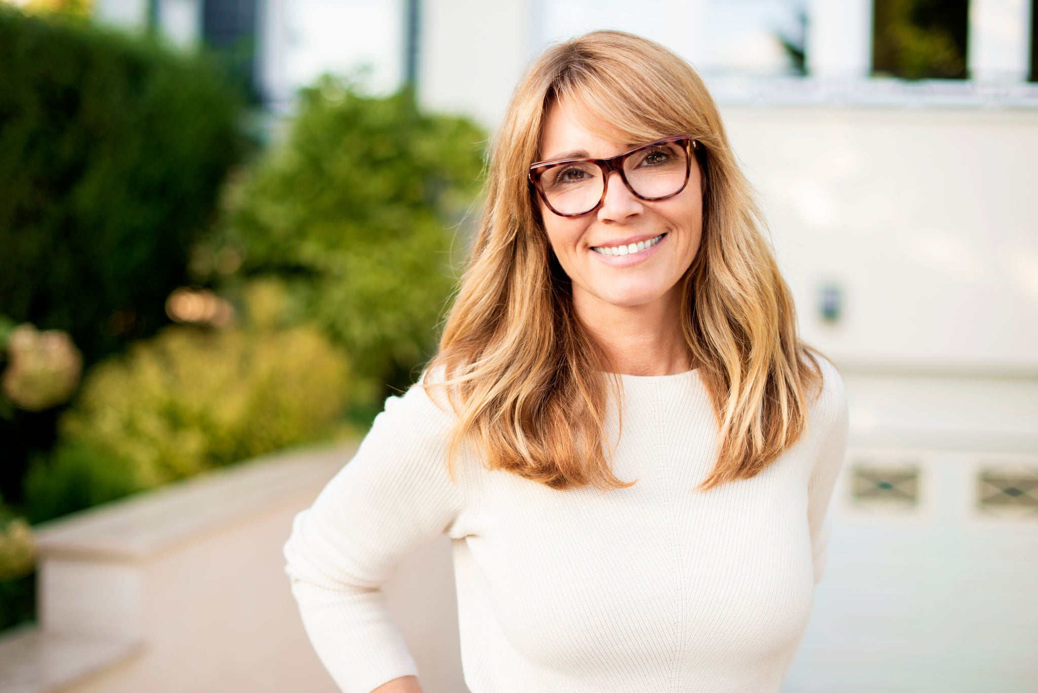 Portrait shot of confident mature woman standing in front yard of home while looking at camera and smiling. 