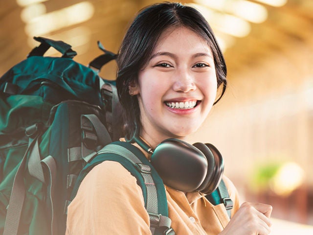 A smiling young woman with a backpack and headphones stands in a bright, spacious environment, suggesting travel or adventure. Resolution: 640x480.