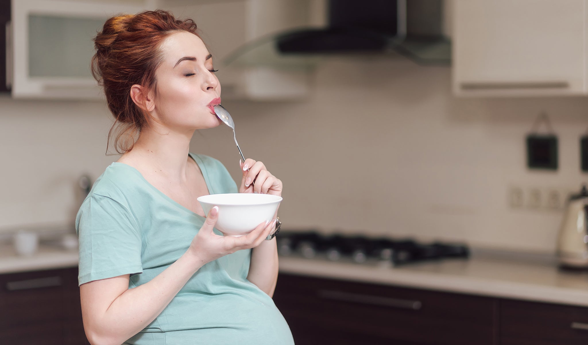 Pregnant female woman with light green shirt eating cereal in kitchen