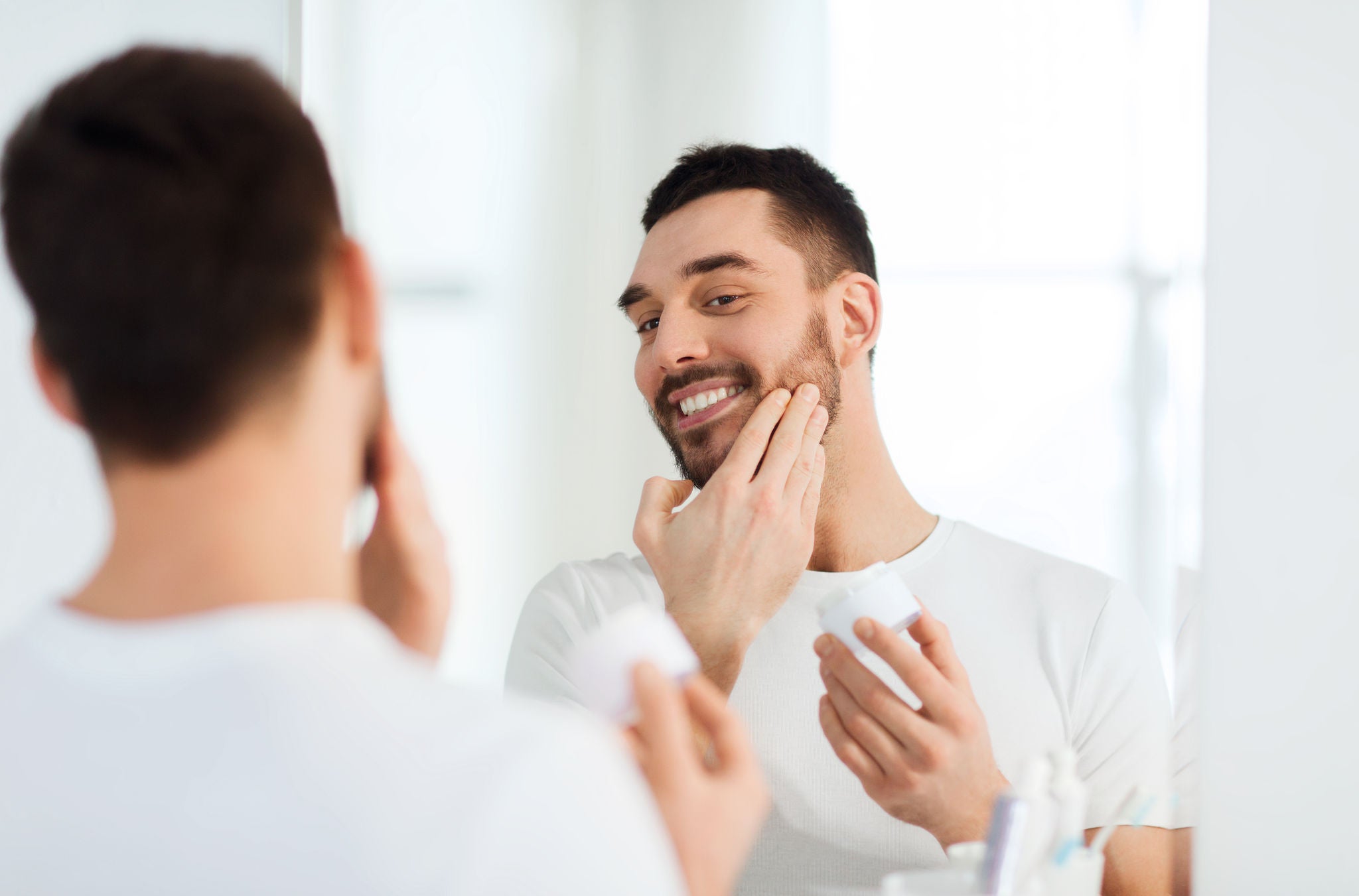 beauty, skin care and people concept - smiling young man applying cream to face and looking to mirror at home bathroom