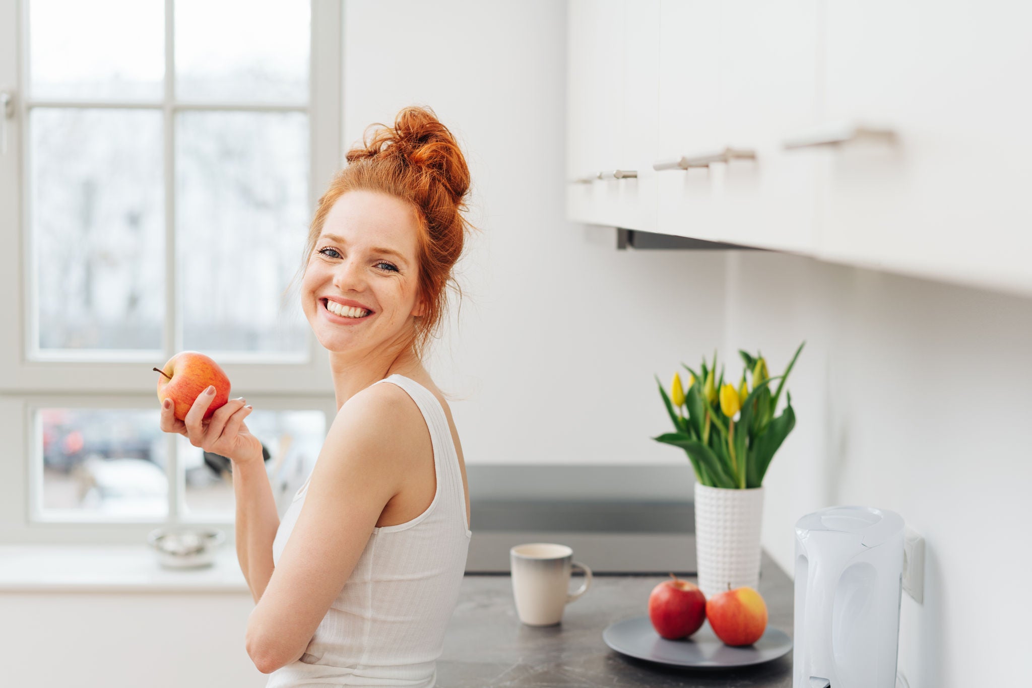 Happy healthy young redhead woman eating fresh fruit holding an apple in her hand as she leans against a kitchen counter grinning at the camera