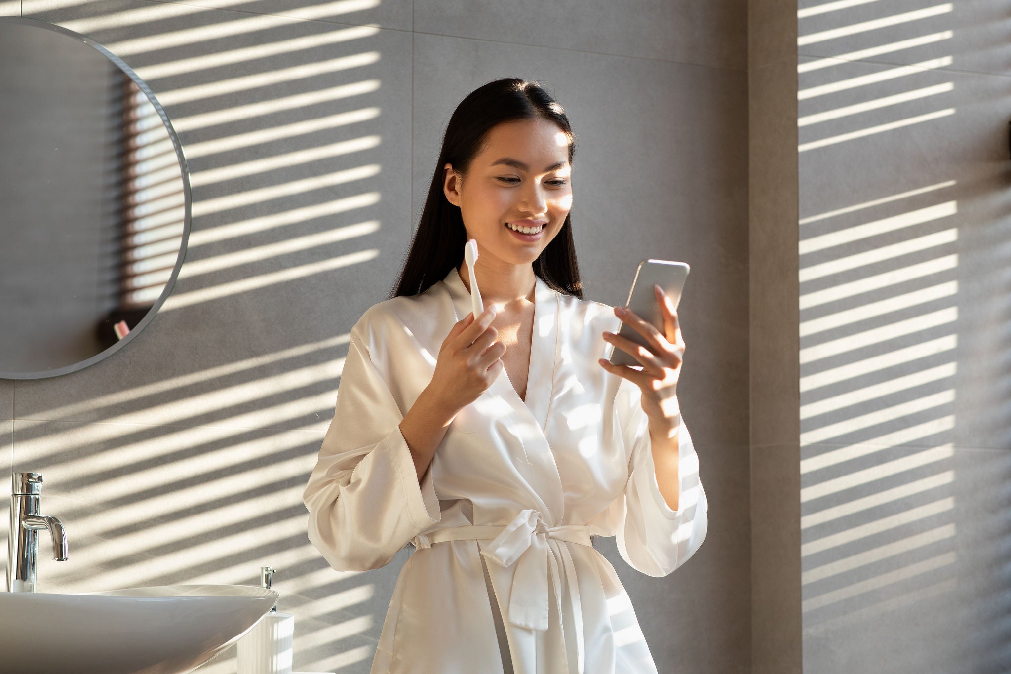 Cheerful long-haired millennial korean lady in silk bathrobe using mobile phone while doing morning routine in bathroom, brushing teeth, looking at smartphone screen and smiling, sun flare