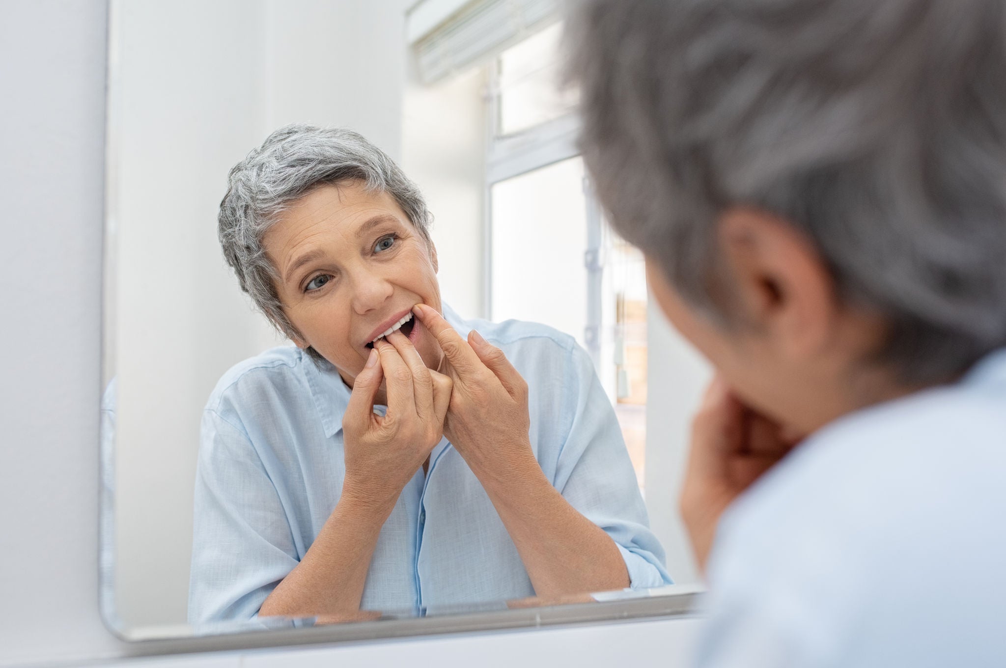 Mature beautiful woman cleaning her teeth with floss in bathroom. Reflection of senior woman in bathroom mirror while cleaning teeth with dental floss. Healthcare and oral hygiene concept.