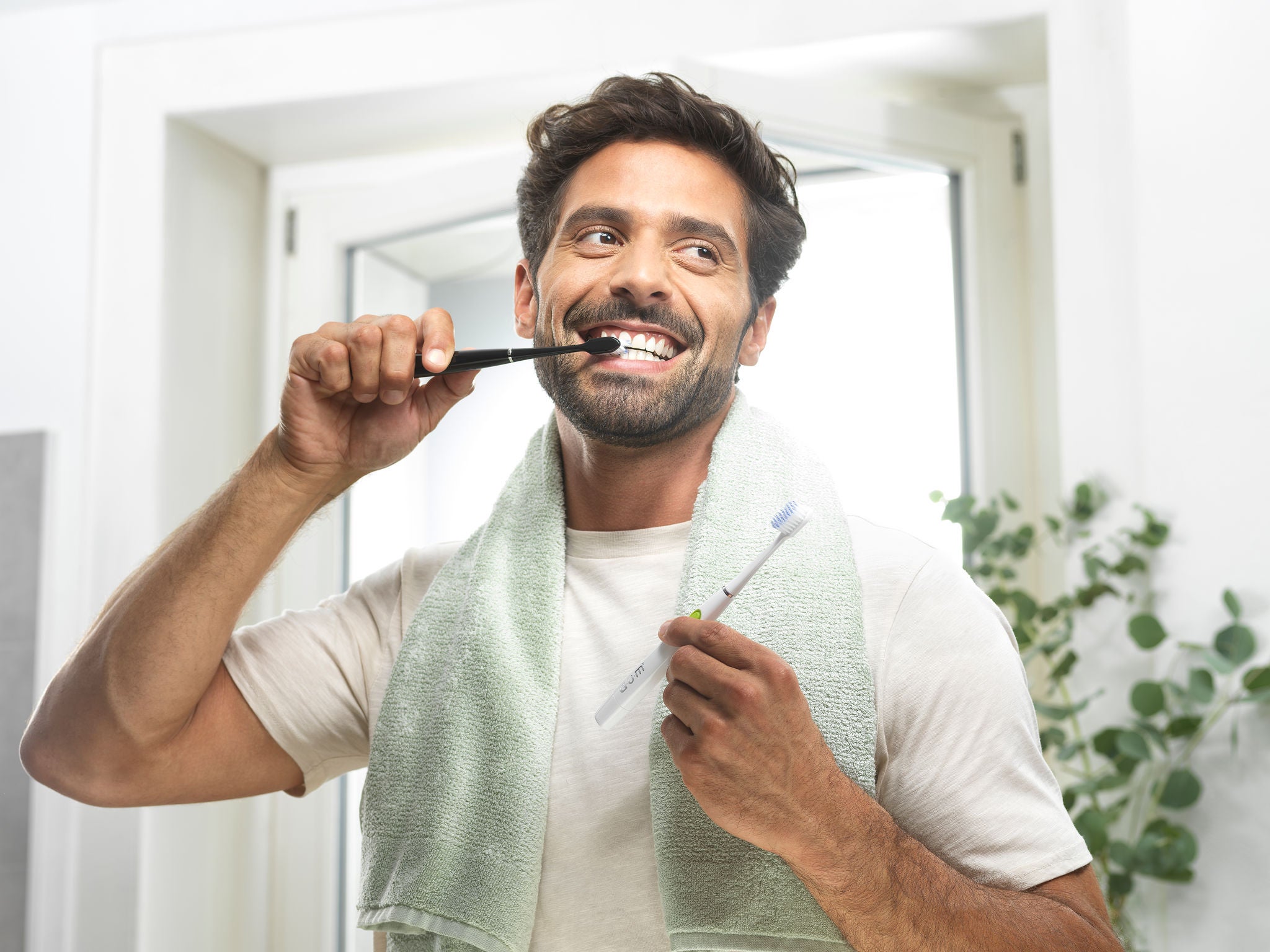 Smiling man is brushing his teeth with the GUM SONIC Toothbrush, the black one, while he's holding the SONIC DAILY toothbrush in white colour
