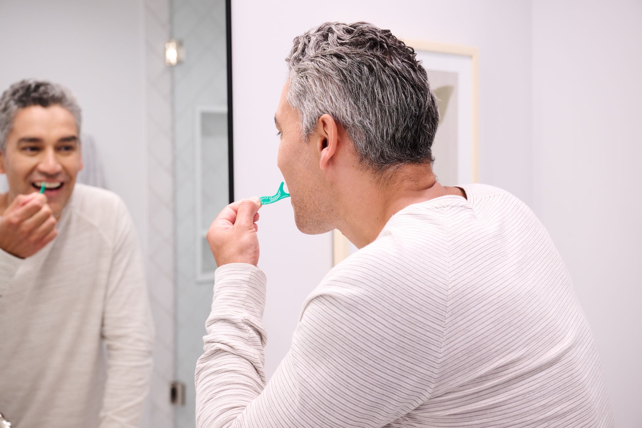 A man uses a green interdental flosser in a bathroom. He's wearing a white shirt, looking in the mirror. Image resolution: 2048x1365.