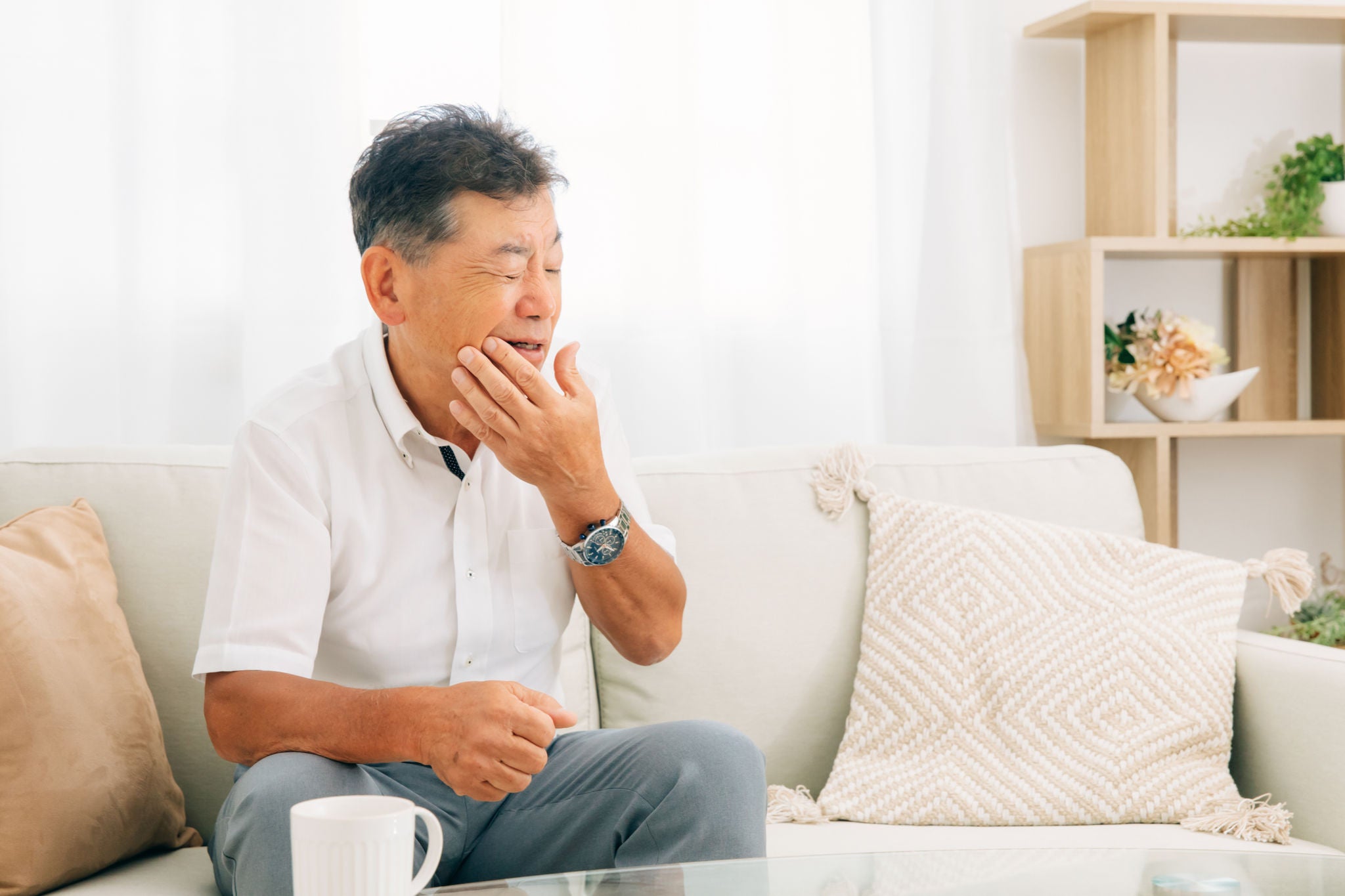 uomo asiatico a sedere sul divano che si tocca la guancia con smorfia di dolore a causa di una nevralgia ai denti