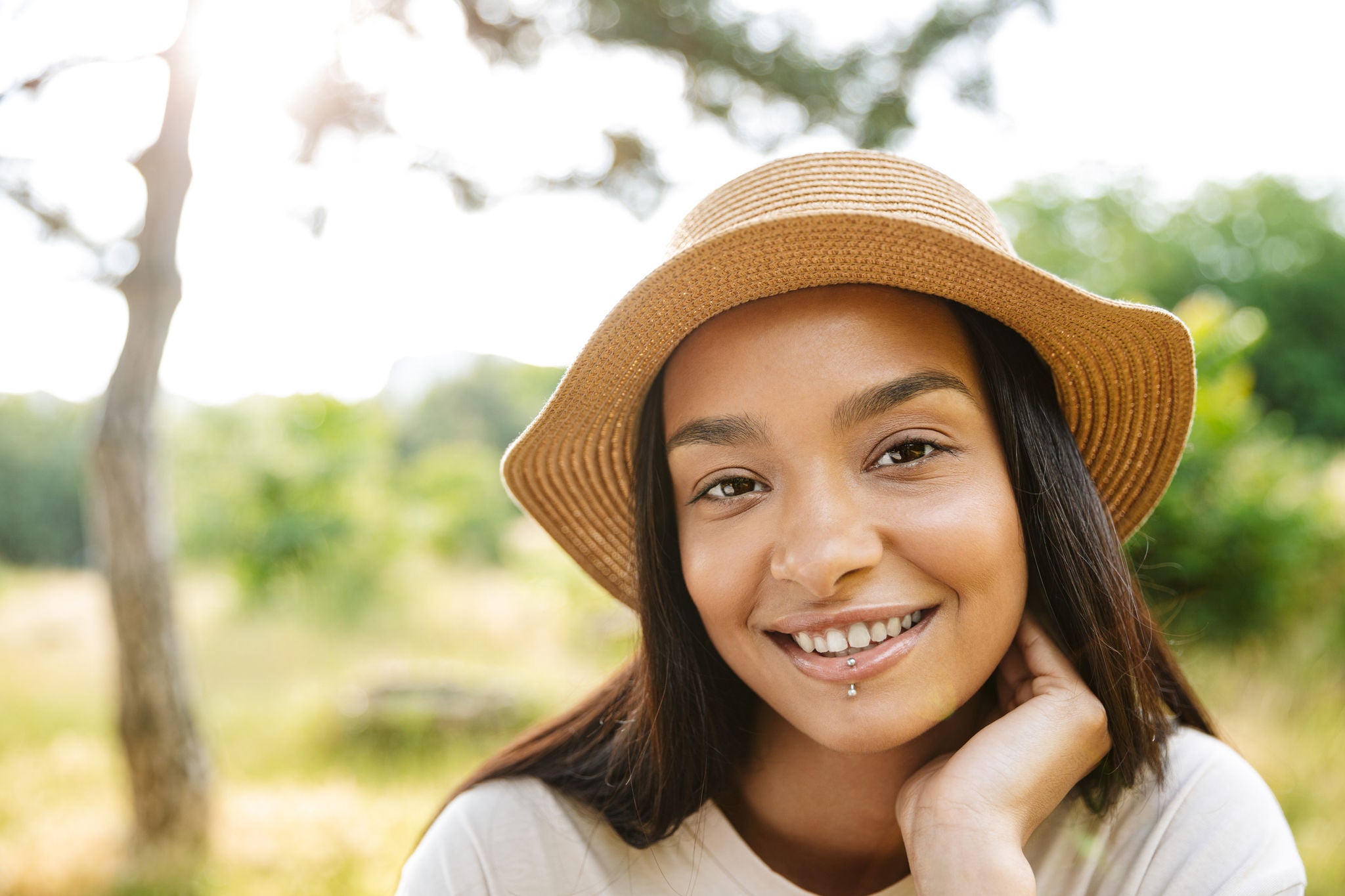 Photo of cheerful woman wearing straw hat and lip piercing smiling at camera while walking in green park