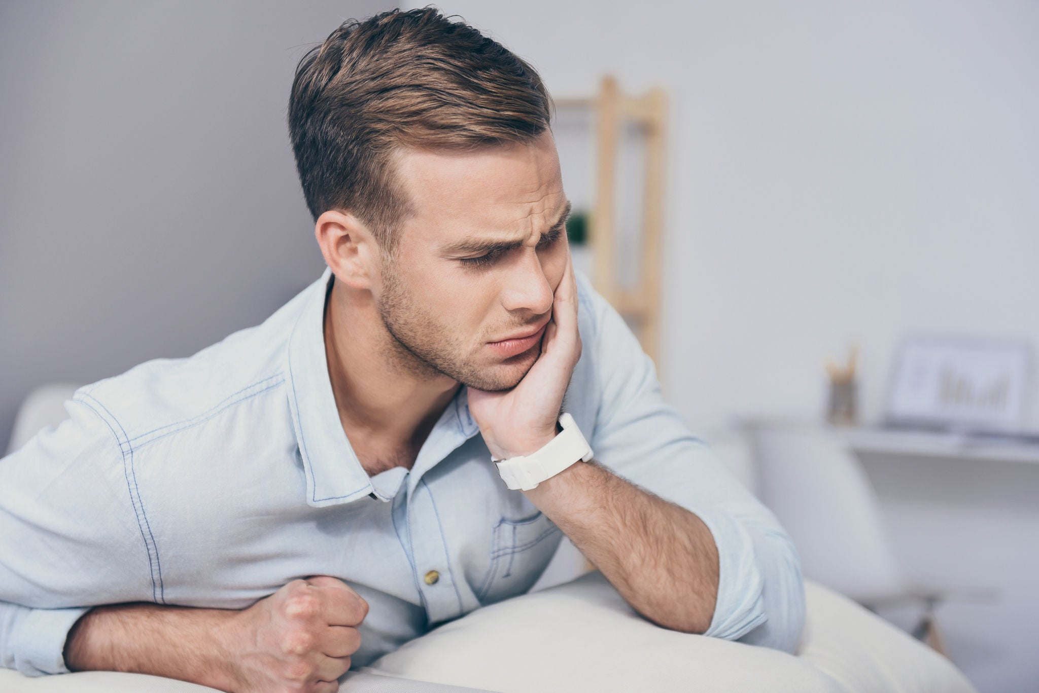 Need a doctor. Handsome young unhappy man closing his eyes and having toothache while sitting in the room.