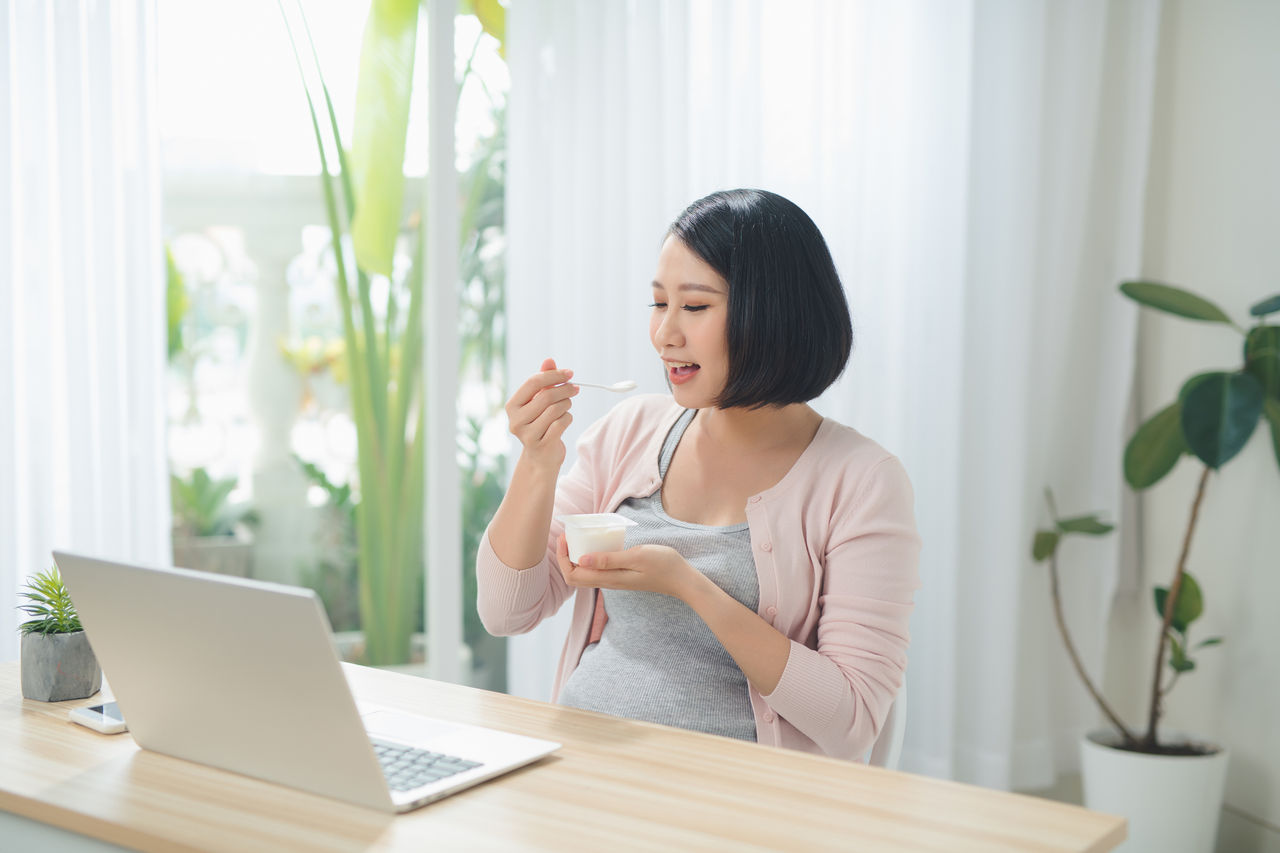Beautiful pregnant business woman is eating yogurt and smiling while sitting at her working place at home