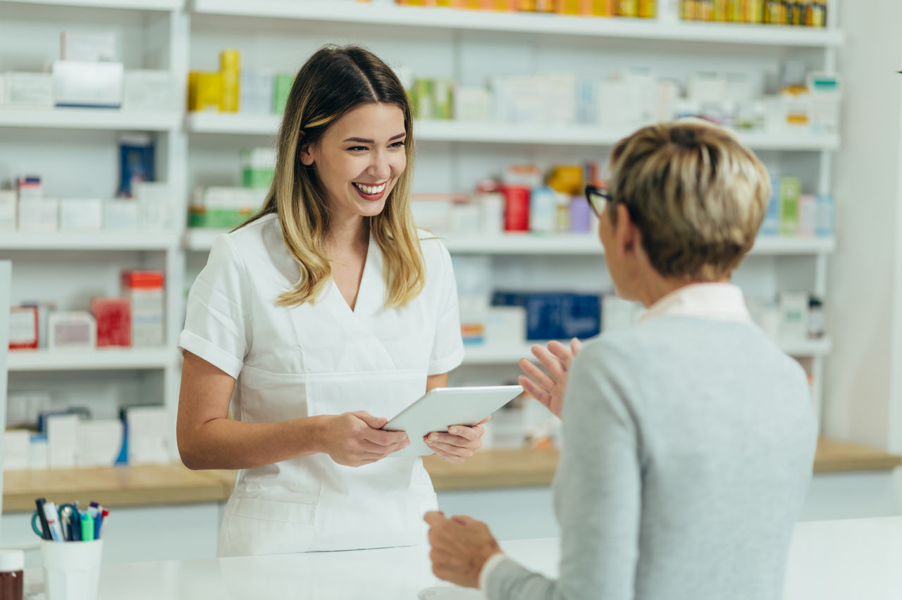 Female pharmacist selling medications at drugstore to a senior woman customer while using a tablet